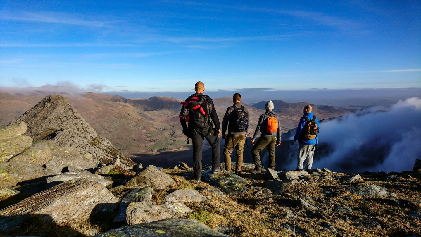 Four backpackers stand on a rocky mountain summit overlooking valleys.